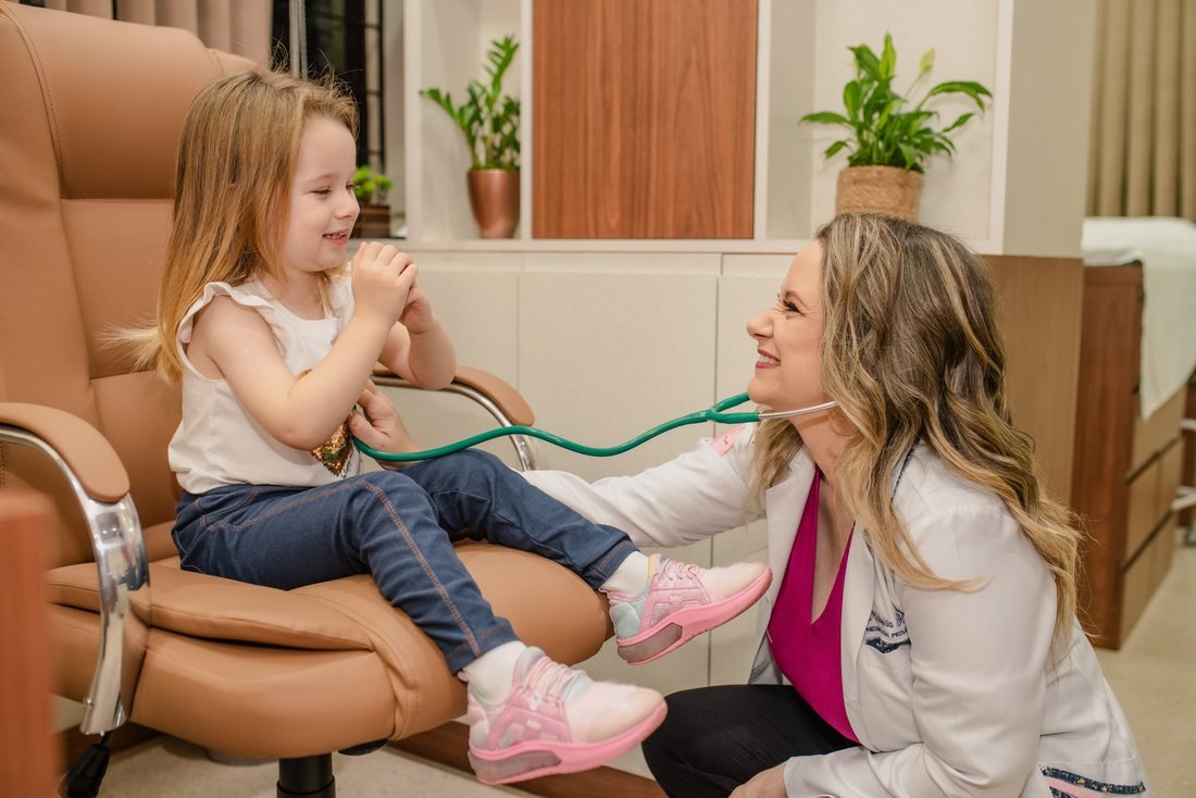 Dra. Valquíria Schroder sorrindo e brincando com uma criança, que segura o estetoscópio dela